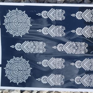 May include: A sheet of white henna stencils on a black background. The stencils feature intricate designs of circles, swirls, and floral patterns. The sheet is labelled "Henna Styles".