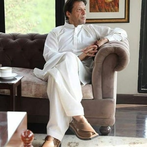 May include: A man wearing a white long-sleeved shirt and white pants sits on a brown couch. He is wearing brown leather sandals. The man is looking to the right of the image.