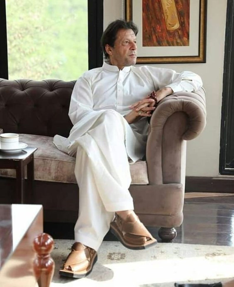 May include: A man wearing a white long-sleeved shirt and white pants sits on a brown couch. He is wearing brown leather sandals. The man is looking to the right of the image.