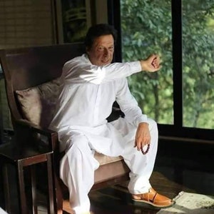 May include: A man wearing a white shalwar kameez sits in a chair with his arm outstretched. He is wearing brown leather sandals and holding a beaded bracelet. The man is sitting in front of a window with a view of green trees.