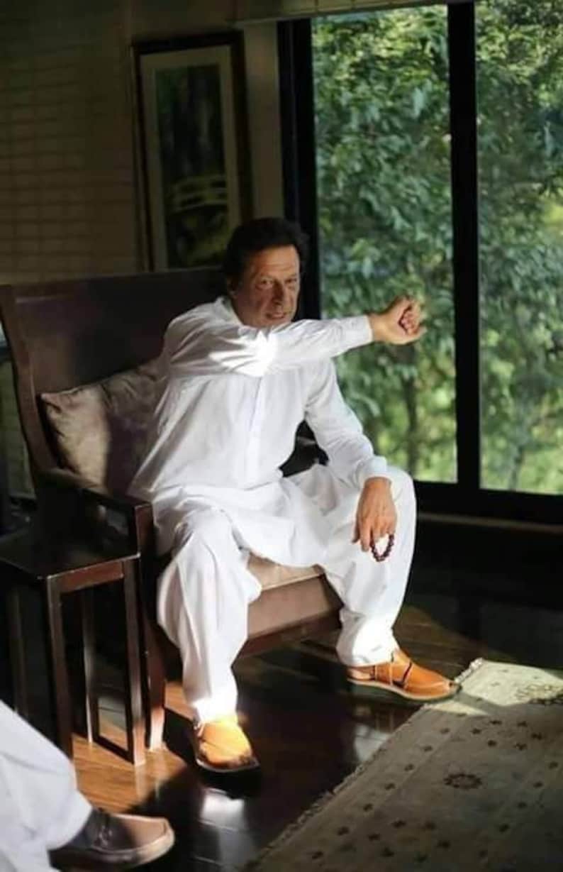 May include: A man wearing a white shalwar kameez sits in a chair with his arm outstretched. He is wearing brown leather sandals and holding a beaded bracelet. The man is sitting in front of a window with a view of green trees.