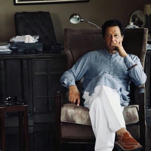 May include: A man wearing a light blue shirt and white pants sits in a brown leather armchair. He is wearing brown leather sandals and has his hand on his chin. The chair is in a room with a dark wood desk and a window in the background.