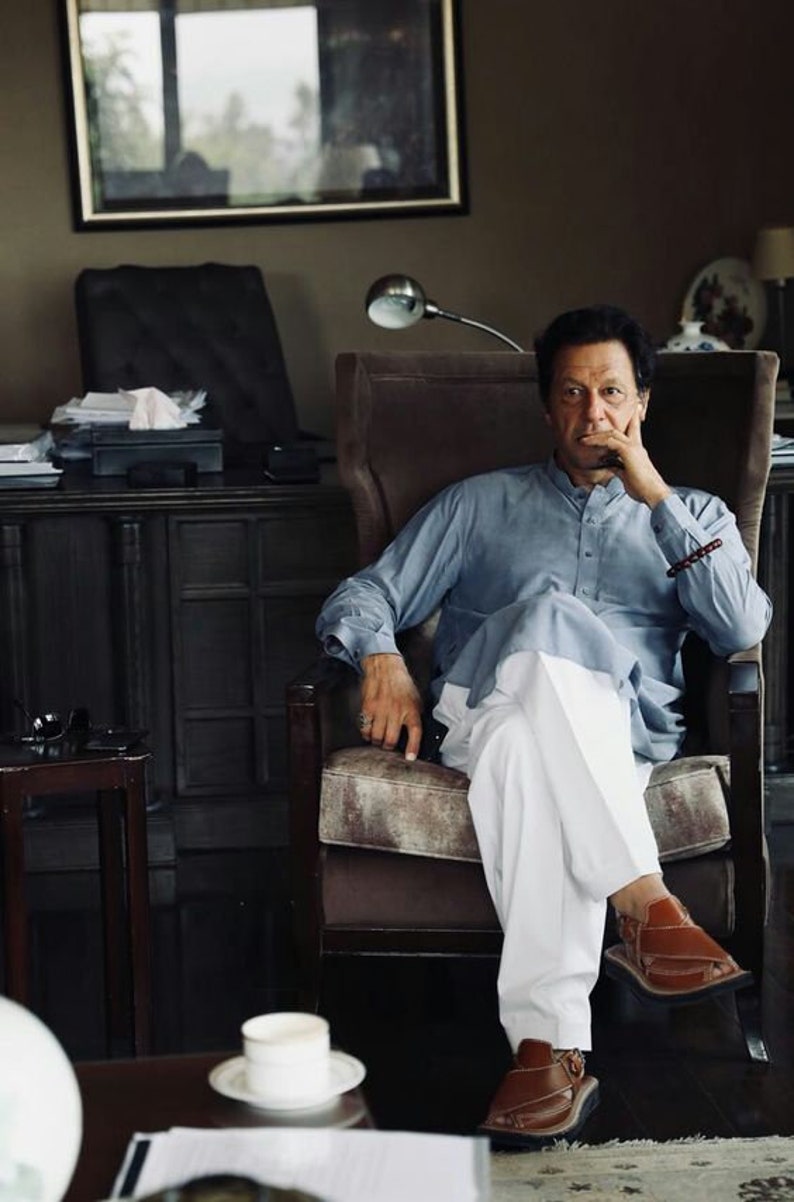 May include: A man wearing a light blue shirt and white pants sits in a brown leather armchair. He is wearing brown leather sandals and has his hand on his chin. The chair is in a room with a dark wood desk and a window in the background.