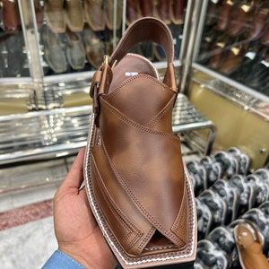 May include: Brown leather sandals with a pointed toe and a white stitching detail around the edge. The sandals have a strap that goes around the ankle and a buckle closure.