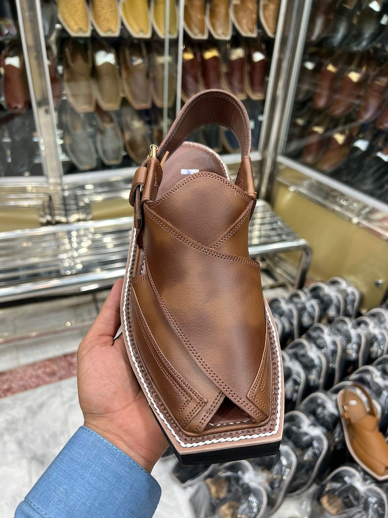 May include: Brown leather sandals with a pointed toe and a white stitching detail around the edge. The sandals have a strap that goes around the ankle and a buckle closure.