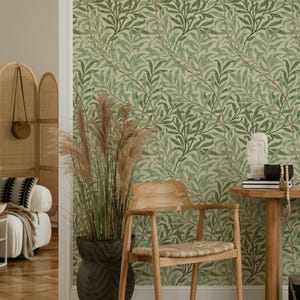 May include: A room with green leaf-patterned wallpaper, a wooden chair with a woven seat, and a round wooden side table. A large black pot with tall, dried plants sits in front of the wallpaper. A white sculpture and books are on the table.