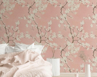 Könnte beinhalten: Tapete mit einem sich wiederholenden Muster aus dunkelbraunen Ästen und cremeweißen Kirschblüten auf einem einfarbigen, altrosa Hintergrund. Das Design bedeckt die Wand hinter einem Bett mit einer hellrosa Decke und weißen Kissen.