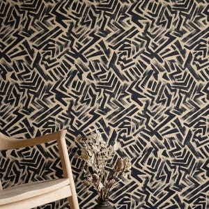 May include: A light-colored wooden rocking chair sits before a wall covered in wallpaper featuring a repeating black brushstroke pattern on a beige background. A dark ceramic vase holds dried flowers next to a small incense holder and a folded cloth.