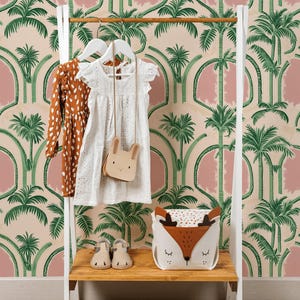 May include: A white and wood clothing rack displays a brown polka-dot dress, a white eyelet dress, and a bunny-shaped purse. A fox-faced storage basket and beige shoes sit on the shelf. The background features a pink and green palm tree wallpaper.