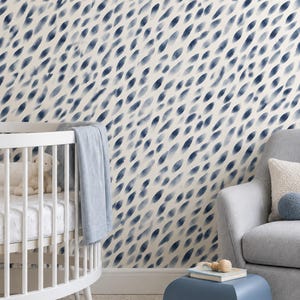 May include: A white wall covered in wallpaper featuring a repeating pattern of dark blue, elongated, brushstroke-like teardrop shapes. The room includes a white round crib, a light gray armchair, and a small blue stool on a circular rug.