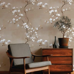 Pode incluir: Um interior de estilo moderno de meados do século com uma poltrona de madeira com almofada cinza, uma cômoda de madeira escura e uma planta em vaso. A parede é adornada com papel de parede floral em tons de bege e branco, criando um ambiente sereno.