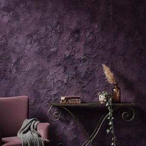 May include: A deep purple textured wall forms the backdrop for a decorative display. A wrought-iron console table holds books, a small potted plant, and a brown glass vase with dried grasses. A matching armchair with a grey throw completes the scene.