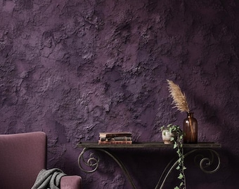 Op de afbeelding: Een dieppaarse, getextureerde muur vormt de achtergrond voor een decoratieve scène. Een smeedijzeren console met boeken, een kleine plant in een pot en een bruine glazen vaas met gedroogde grassen. Een bijpassende fauteuil met een grijze plaid maakt het geheel compleet.