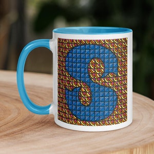 May include: A white ceramic mug with a blue rim and a colorful pattern featuring the letter "S" in blue. The pattern is made up of small squares and circles in shades of orange, yellow, and pink.
