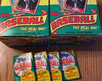 May include: Two boxes of 1987 Topps Baseball bubble gum cards with baseball cards on top. The boxes are yellow and green with the words "BASEBALL" and "THE REAL ONE" printed on them. Four packs of baseball cards are in front.