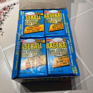 Puede incluir: Una caja de cromos de pegatinas con el logotipo de béisbol Fleer de 1987. Las cajas azules tienen texto blanco y amarillo. La caja dice "Hey Kids! Collect the expanded 1987 series of Limited Edition Cards & Logos on Box Bottom."