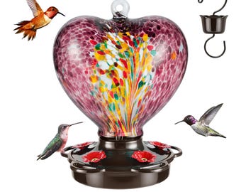 May include: A heart-shaped glass hummingbird feeder with a colorful, speckled design. The feeder has four feeding ports and a metal hook for hanging. Three hummingbirds are flying around the feeder.