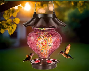 May include: A decorative hummingbird feeder with a heart-shaped, speckled glass reservoir in shades of pink, red, and yellow. The feeder has a dark brown top and base, with two hummingbirds nearby. Text at the top reads "Lgnite Your Garden's Glow."