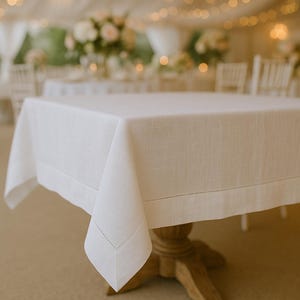 Puede incluir: Un mantel cuadrado de color blanco con una textura tejida visible y un detalle de borde con dobladillo calado a lo largo de los lados. La tela cae sobre las esquinas de una mesa de madera, mostrando un color blanco sólido y un acabado estructurado.