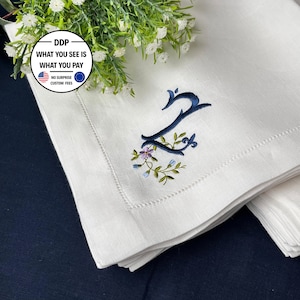 Peut inclure: Une serviette en lin blanc ornée d'une lettre Z brodée en bleu foncé, agrémentée de petites feuilles vertes et de minuscules fleurs violettes et bleues. La serviette présente un bord à jour et est pliée, avec un bouquet de fleurs blanches.