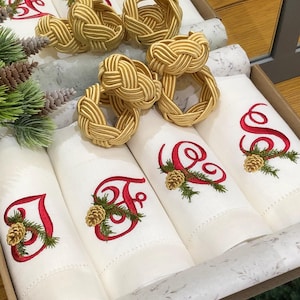 Puede incluir: Un juego de servilletas de tela blanca con letras bordadas en rojo y motivos de piñas, junto con servilleteros de cuerda trenzada dorada. Las servilletas están dispuestas en una caja junto a ramas de pino verdes y pequeñas piñas.
