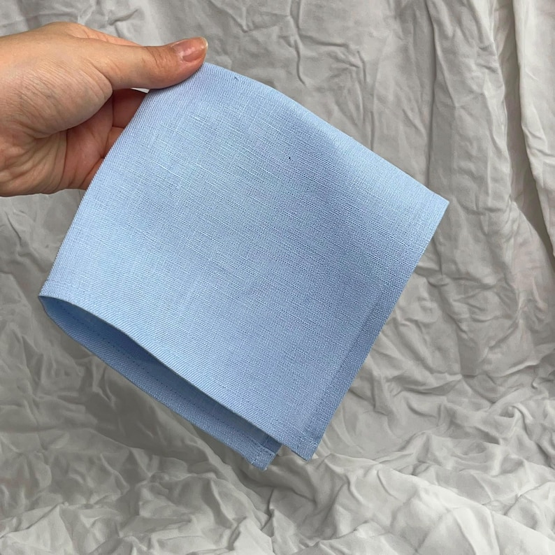 K&ouml;nnte beinhalten: Ein quadratisches hellblaues Taschentuch aus einem gewebten Stoff mit sichtbarer Textur. Das Tuch ist zu einem Dreieck gefaltet und weist saubere, gerade Kanten entlang des Umfangs auf, die eine einheitliche, pastellblaue Farbe zeigen.