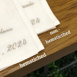 K&ouml;nnte beinhalten: Zwei cremewei&szlig;e Stoffservietten mit gesticktem Text, einschlie&szlig;lich der Jahreszahl 2024. Eine Serviette hat einen Hohlsaum, die andere nicht. Der Text 'hemstiched' und 'non-hemsticthed' ist zum Vergleich auf das Bild gedruckt.