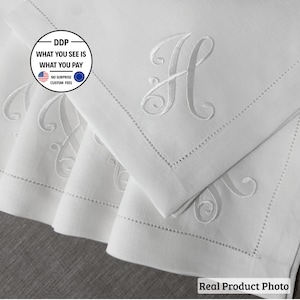 May include: A set of white linen napkins featuring an embroidered script letter H in the center. Each napkin has a decorative hemstitch border detail along the edges. The fabric is a crisp white, showcasing the tonal embroidery work clearly.