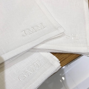 K&ouml;nnte beinhalten: Drei cremewei&szlig;e Leinenservietten mit gestickten Monogrammen. Eine Serviette zeigt die Buchstaben JMM, eine weitere die Buchstaben SH und die dritte Serviette die Buchstaben GMT. Jedes Monogramm ist mit passendem cremewei&szlig;em Garn gestickt.