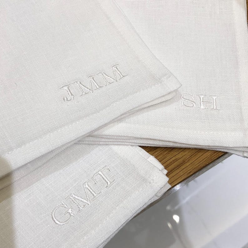 K&ouml;nnte beinhalten: Drei cremewei&szlig;e Leinenservietten mit gestickten Monogrammen. Eine Serviette zeigt die Buchstaben JMM, eine weitere die Buchstaben SH und die dritte Serviette die Buchstaben GMT. Jedes Monogramm ist mit passendem cremewei&szlig;em Garn gestickt.