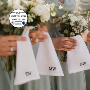 K&ouml;nnte beinhalten: Drei wei&szlig;e Stofftaschent&uuml;cher, jedes mit einem einzigartigen, in schwarzem Garn gestickten zweibuchstabigen Monogramm. Die Monogramme lauten TS, MW und SW. Jedes Taschentuch wird neben einem Strau&szlig; aus wei&szlig;en Rosen und gr&uuml;nem Eukalyptus gehalten.