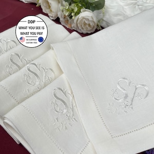 Puede incluir: Un juego de servilletas de lino blanco con un monograma bordado de las letras SP en una fuente cursiva decorativa. Cada servilleta tiene un borde de vainica y está doblada para mostrar el bordado. Se observa un logotipo circular con texto DDP.