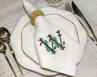 May include: A white cloth napkin featuring an embroidered green letter W intertwined with green leaves and small red berries. The napkin is secured with a gold-colored rope-style napkin ring and placed on a white plate with a thin gold rim.