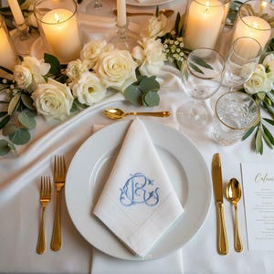 Puede incluir: Elegante mesa con rosas blancas, velas y cubiertos dorados. Un plato blanco sostiene una servilleta doblada bordada con iniciales azules. Copas de vino y una tarjeta de menú completan el arreglo formal.