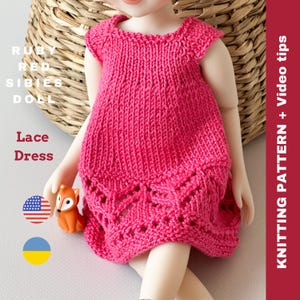 Puede incluir: Un vestido de encaje de punto rosa para una muñeca. El vestido tiene un corpiño sencillo y un borde de encaje alrededor del dobladillo. El vestido lo lleva una muñeca sentada en una cesta de mimbre.