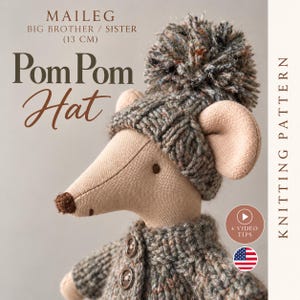 May include: A beige fabric mouse toy wearing a dark gray knit pom pom hat and a matching button-up cardigan. Text on the image reads: MAILEG BIG BROTHER / SISTER (13 CM) Pom Pom Hat KNITTING PATTERN + VIDEO TIPS with a US flag icon.