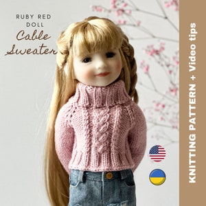 Op de afbeelding: Een roze kabelgebreide trui voor een Ruby Red pop. De trui heeft een rolkraag en wordt getoond op een pop die blauwe jeans draagt. De afbeelding bevat de tekst "Ruby Red Doll Cable Sweater" en de vlaggen van de Verenigde Staten en Oekraïne.