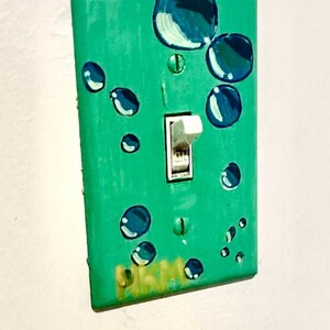 May include: A light switch cover painted teal green with a design of blue circles. The switch is white and is in the off position.