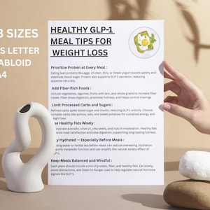 May include: A white paper with the title "HEALTHY GLP-1 MEAL TIPS FOR WEIGHT LOSS" and meal tips. A white ceramic vase and two stacked stones are in the foreground. The paper is held by a hand.