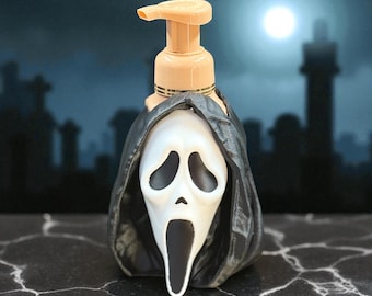 Puede incluir: Un dispensador de jabón beige con un diseño de máscara de Ghostface en blanco y negro. El dispensador tiene una bomba beige y está sobre un fondo oscuro y espeluznante con una luna llena. La máscara tiene grandes cuencas de los ojos y una boca abierta.