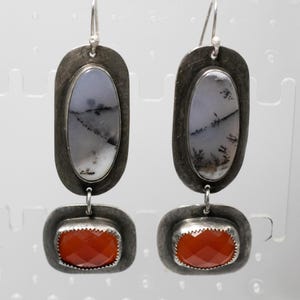 May include: A pair of silver dangle earrings. Each earring has an oval, pale stone with black and grey patterns, and a rectangular, faceted orange stone. The earrings hang from silver hooks. The stones are set in silver frames.