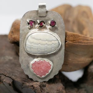 May include: A silver pendant with a rectangular shape, featuring a white and gray striped oval stone, a pink heart-shaped stone, and three small red stones. The pendant is displayed on a wooden surface.