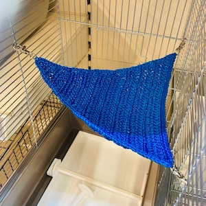 May include: A blue knitted hammock for small pets, hanging inside a wire cage. The hammock is attached to the cage with two metal clips.