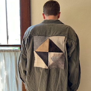 May include: A green utility shirt with a patchwork square on the back. The patchwork is made of different fabrics in shades of brown, black, and white.