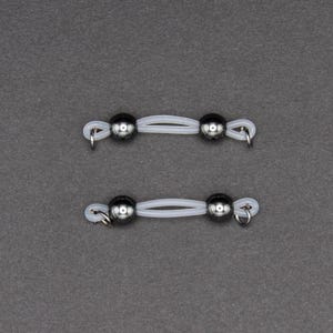 May include: Two sets of body jewelry. Each set features two shiny, dark gray metal balls connected by a thin, white, flexible band. Small silver rings are attached to the ends of the bands.