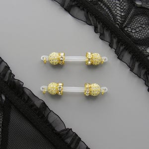 May include: Four gold-colored nipple rings with clear bars and sparkling accents are displayed on a gray surface. Black lace fabric with a ruffled edge frames the top and bottom of the image.