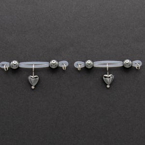 May include: Two nipple rings with a clear band, silver beads, and a heart charm. The jewelry is displayed on a dark gray background. Each ring has a silver heart pendant hanging from the center.