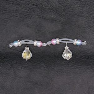 May include: A delicate, clear elastic bracelet adorned with iridescent beads and sparkling rhinestone accents. Two teardrop-shaped crystal pendants dangle from the bracelet, creating a subtle, elegant design. The bracelet is set against a dark, textured background.