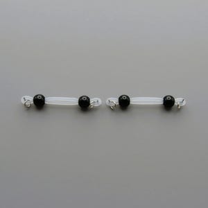 May include: Two pairs of clear, flexible nipple clamps, each with two black beads and small silver-colored rings. The clamps are designed to be worn on the nipples.