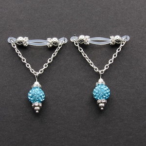 May include: A pair of nipple jewelry with a clear silicone band, silver-colored chain, and a blue, crystal-covered pendant. The jewelry is designed to be worn as body adornment.